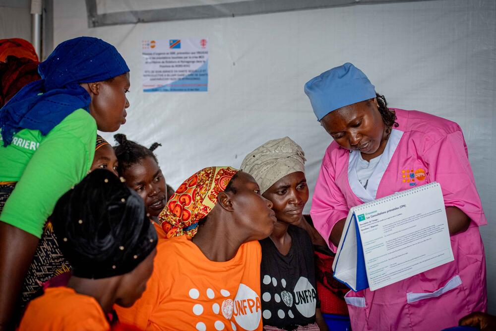 A group of women look at a piece of paper being held up by a nurse clad in a UNFPA vest.