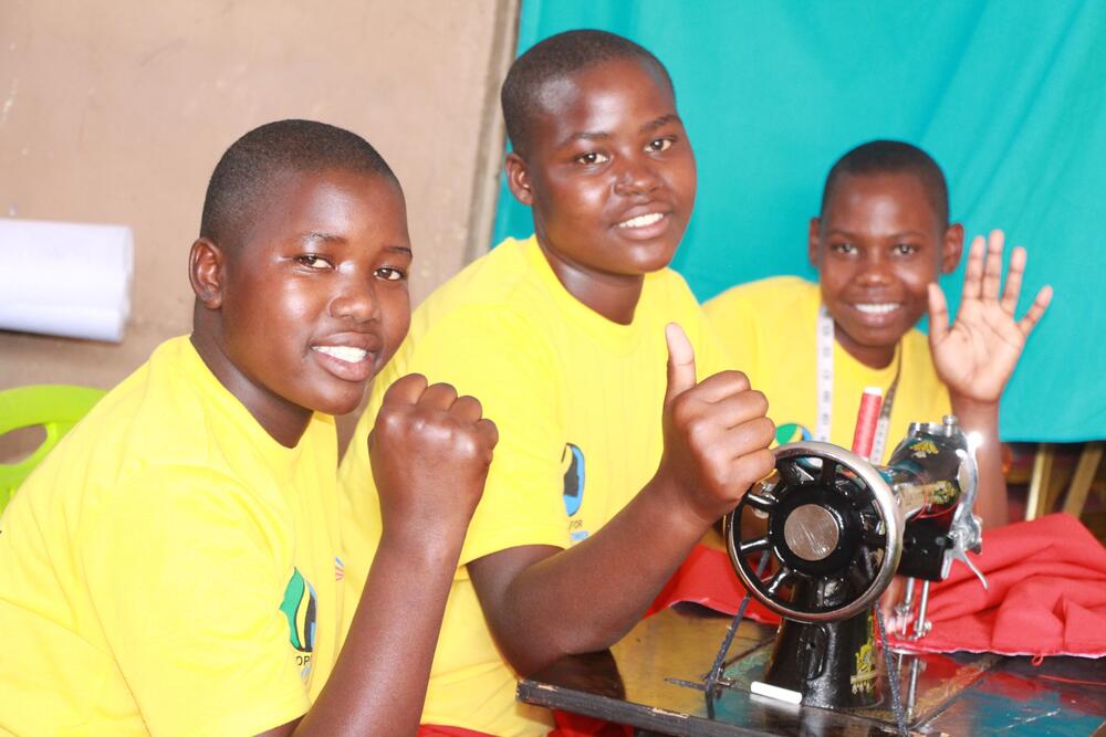 Three girls dressed in school clothes smille at the camera while sitting at a desk with a sewing machine.