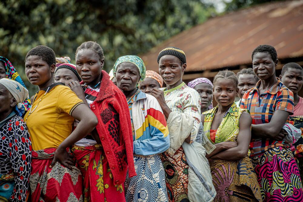 A group of women stand close together in a line. Their colorful clothes stand in contrast with their solemn faces.