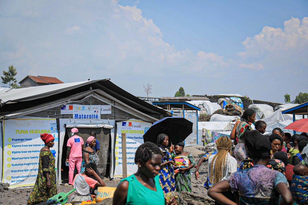 In front of a mobile health clinic a buisy group of women of all ages walk and talk to each other. A nurse is seen going into the clinic.