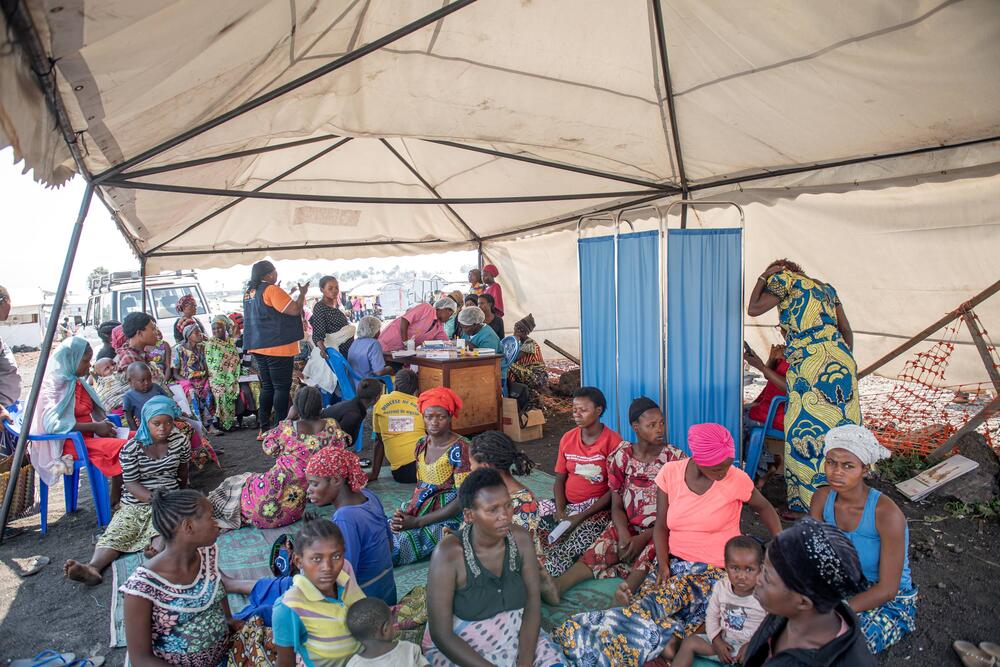 A group of women are huddled together while they wait below a large tent to be seen by a nurse.