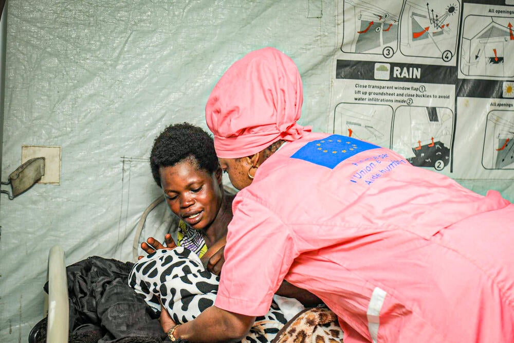 A young woman is being handed her daughter by a nuse clad in pink coveralls.