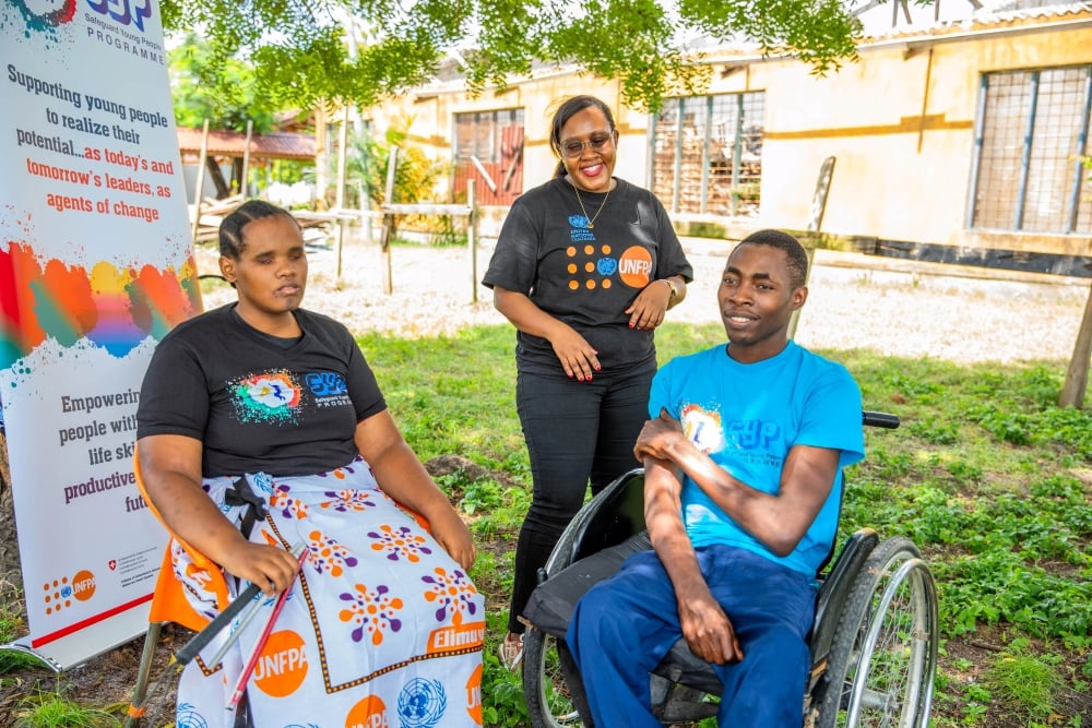 On a brightly lit garder, under the trees, three people look happy while they chat. One of them sits on a wheelchair. 