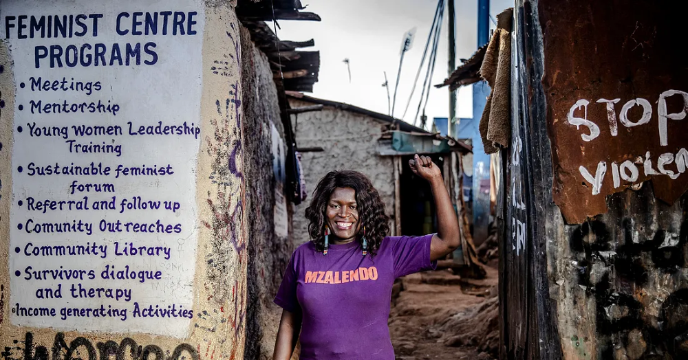 woman stands in front of a feminist center with her fist raised up, smiling at the camera.