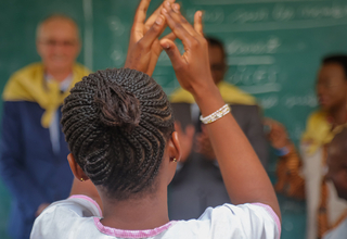 A young girl raises her hands at a classroom setting, in front of her we see teachers and a big blackboard. 