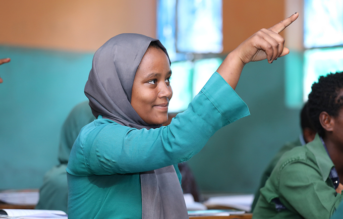 A young girl sitting in a classroom smiles while raising her hand.