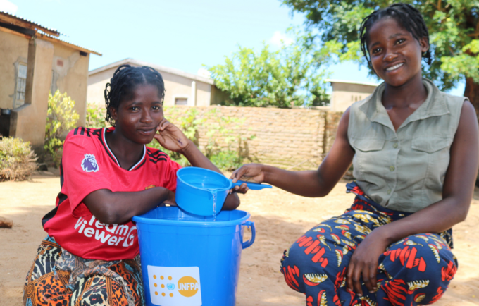 Atupele and her sister Shanil at home next to a plastic tub containing water with the UNFPA logo.