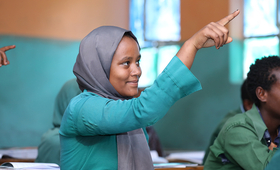 A young girl sitting in a classroom smiles while raising her hand.