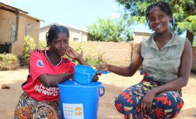 Atupele and her sister Shanil at home next to a plastic tub containing water with the UNFPA logo.