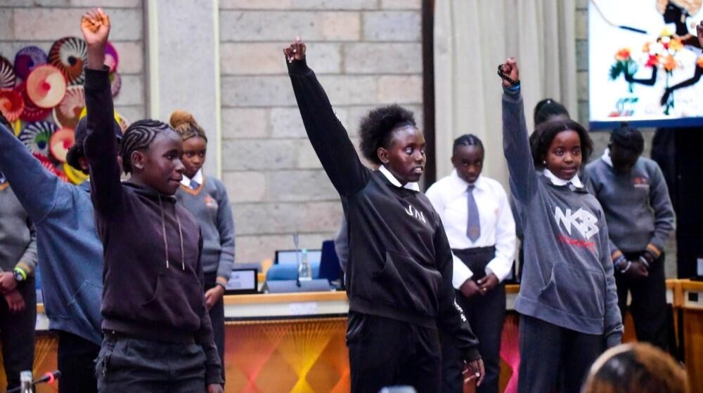 A group of girls raise their fists during a presentation in a closed room.