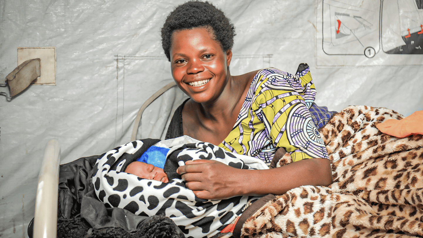 Francine Toyata is laying on a cot, holding her baby and smiling to the camera.