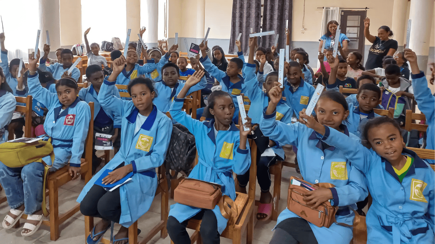 A group of girls raise their hands while sitting at a classroom. They smile to the camera energetically.