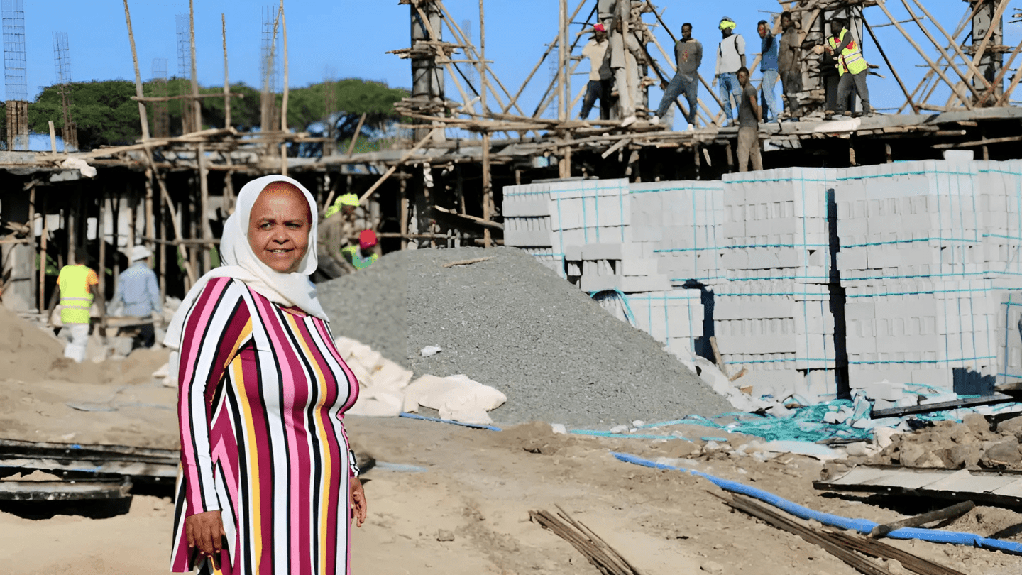 A woman smiles to the camera, in the background a construction site shows a grey block building.