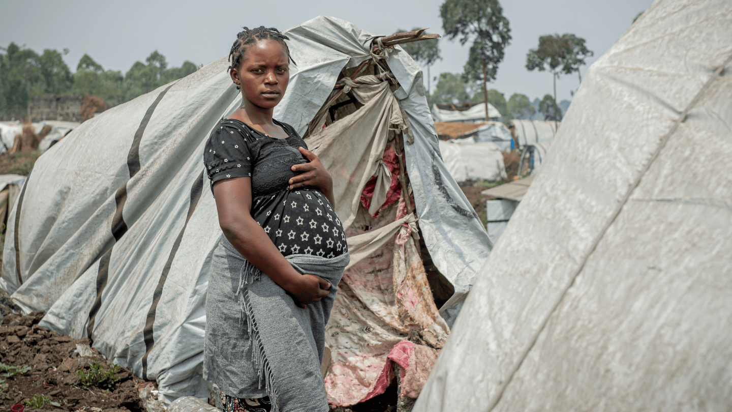 Safi Joséphine, 23, looks seriously at the camera. Tents surround her as she stands in the middle of the displacement camp.