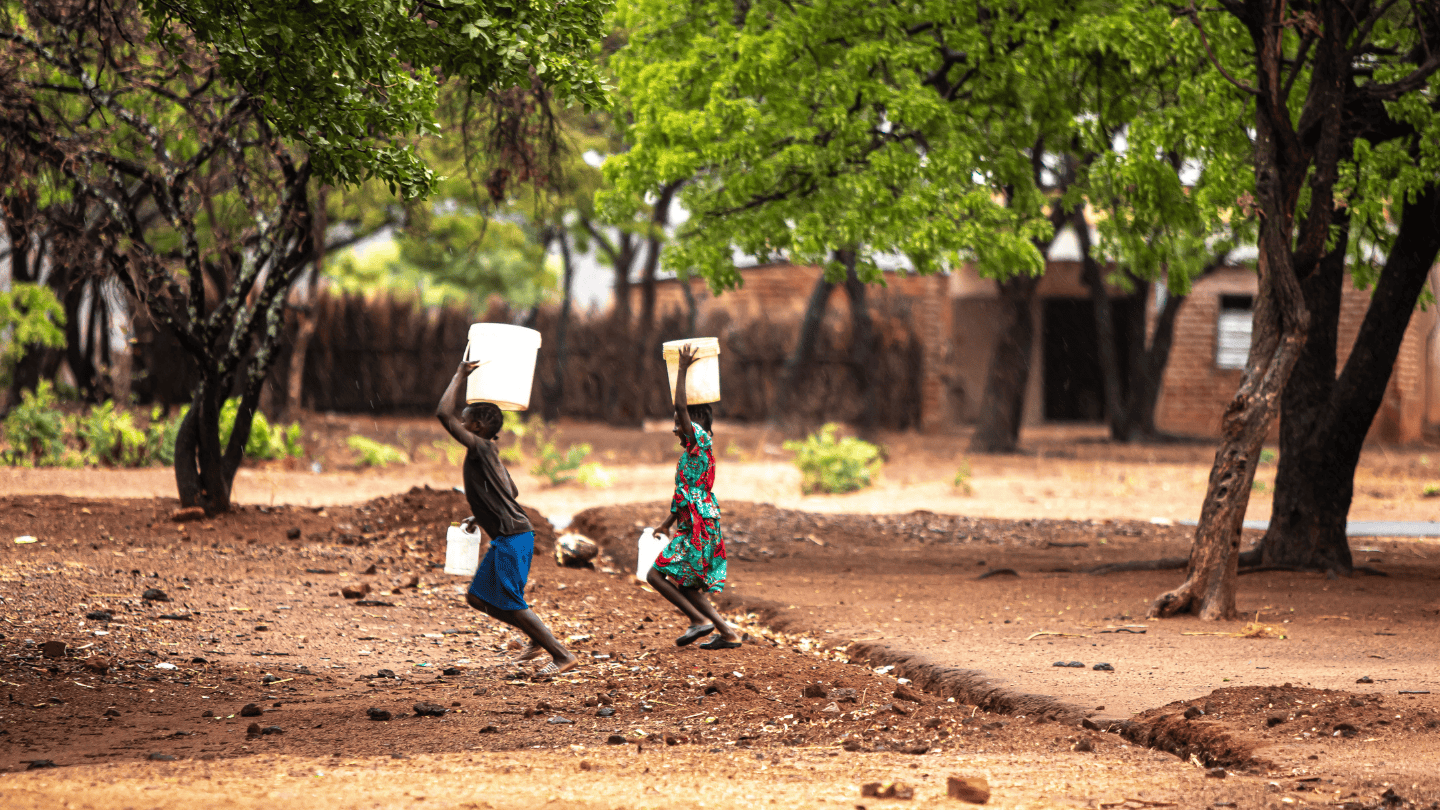 Two young girls walk under trees, the buckets in their hands and heads signal they are walking to fetch water.