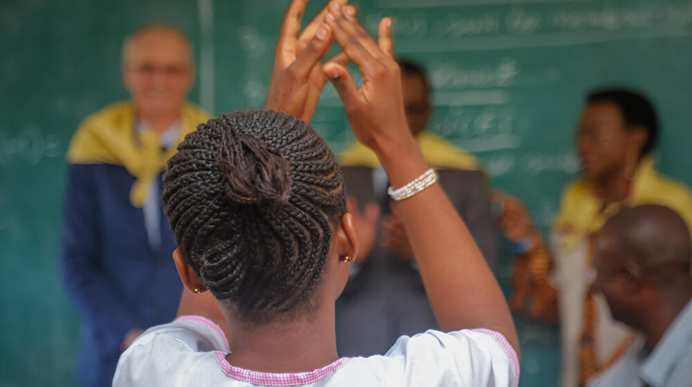 A young girl raises her hands at a classroom setting, in front of her we see teachers and a big blackboard. 